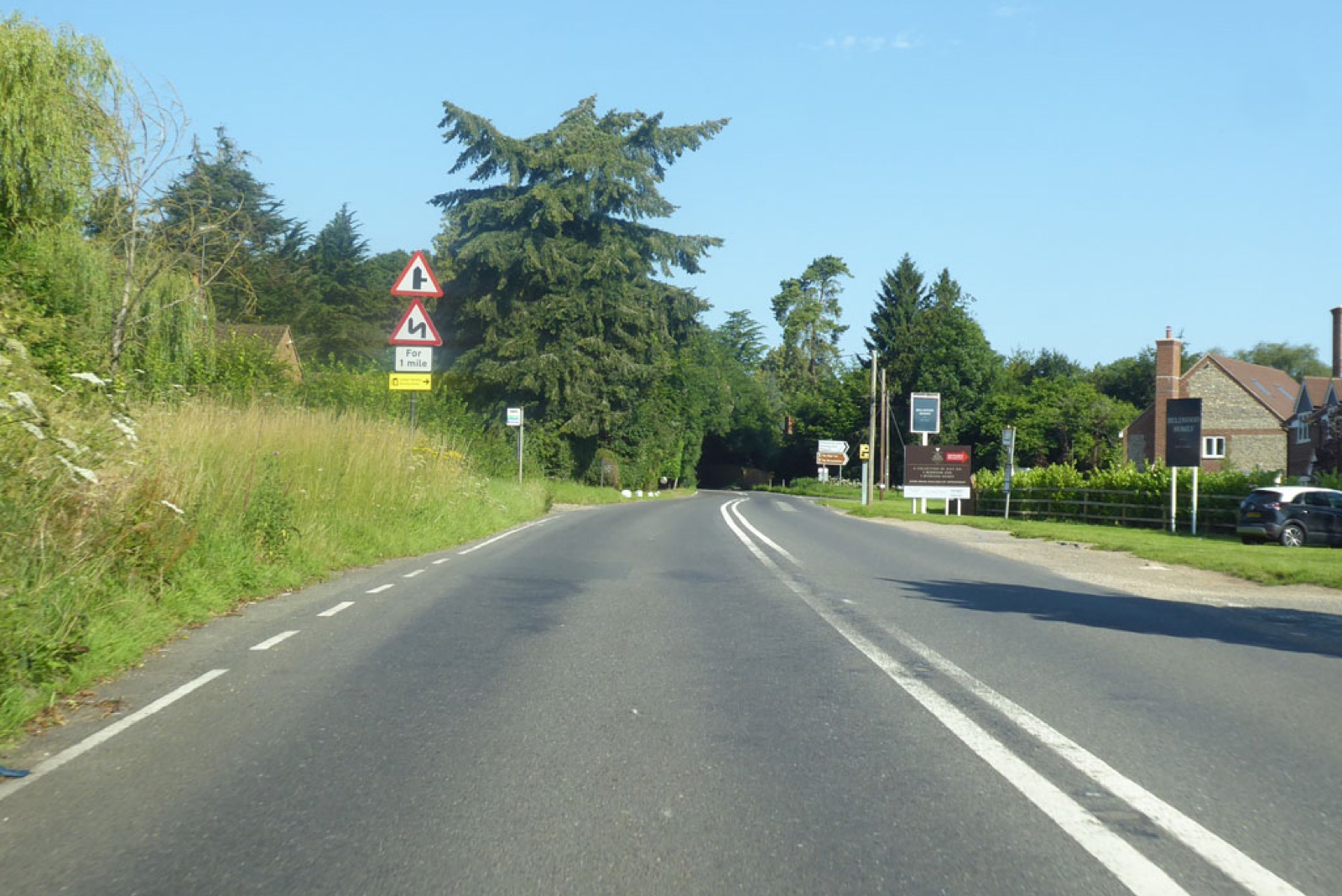The A4074 Reading Road at Cane End, a two-lane country road lined with trees and verges, with bend warning signs on the left and houses set back on the right.