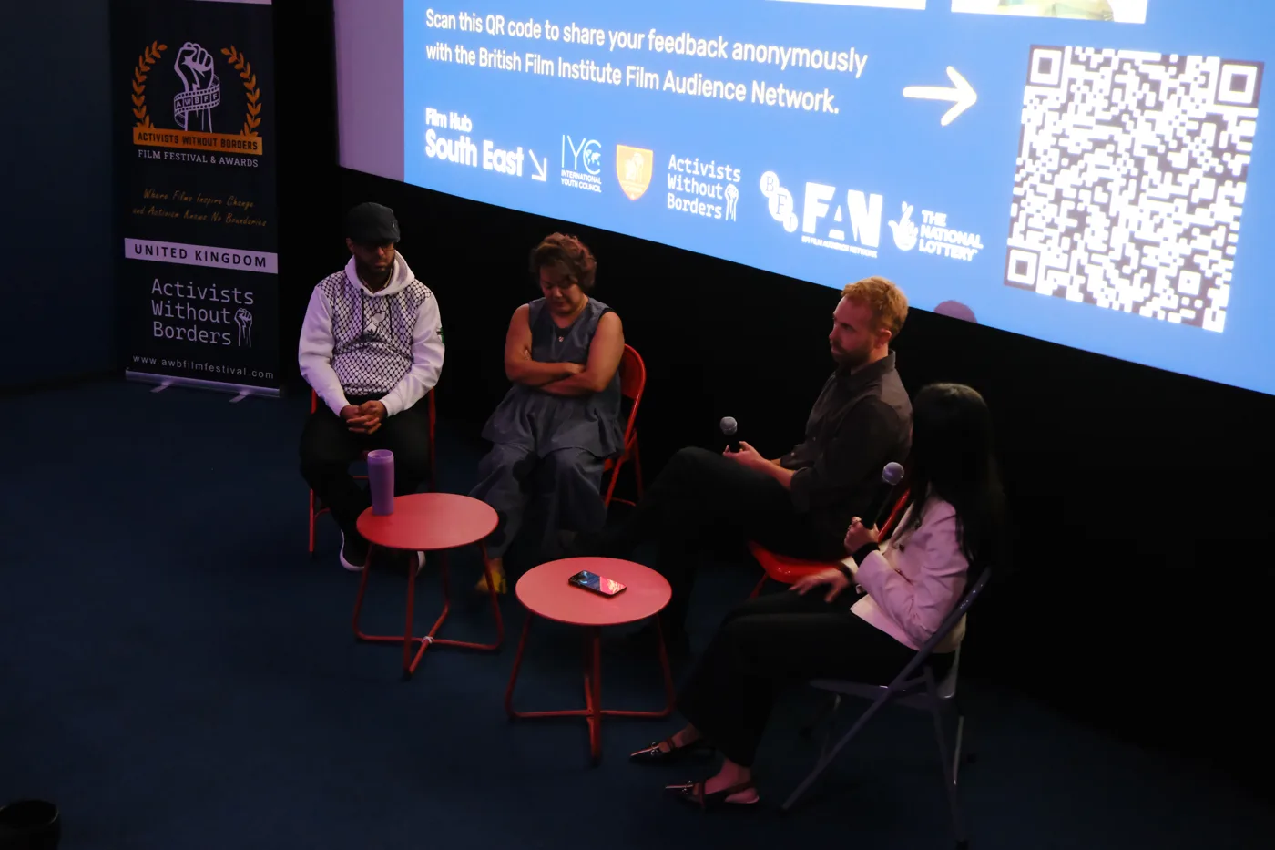 Four people seated on red chairs in front of a cinema screen, two holding microphones, beside an Activists Without Borders banner
