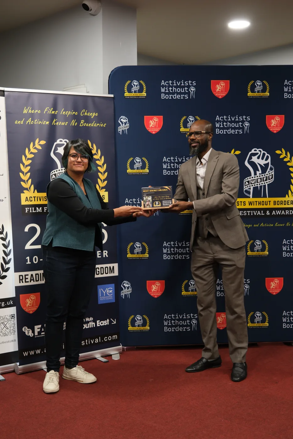 A man in a grey suit hands a Best Short Drama award plaque to a woman in a green waistcoat in front of the festival backdrop