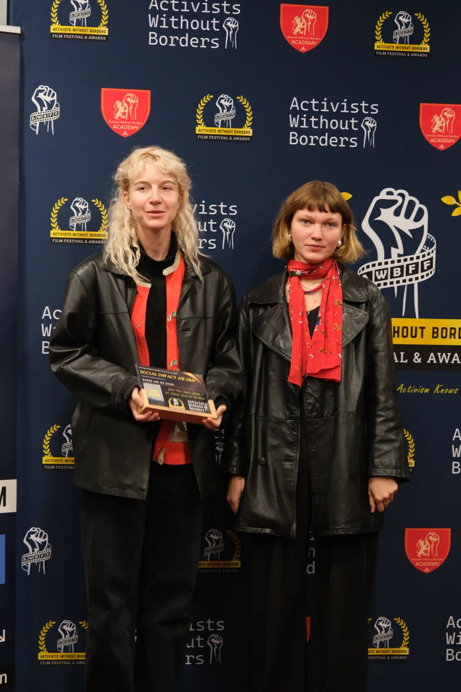 Two young women in black leather jackets, one holding a Social Impact Award plaque, in front of the festival backdrop