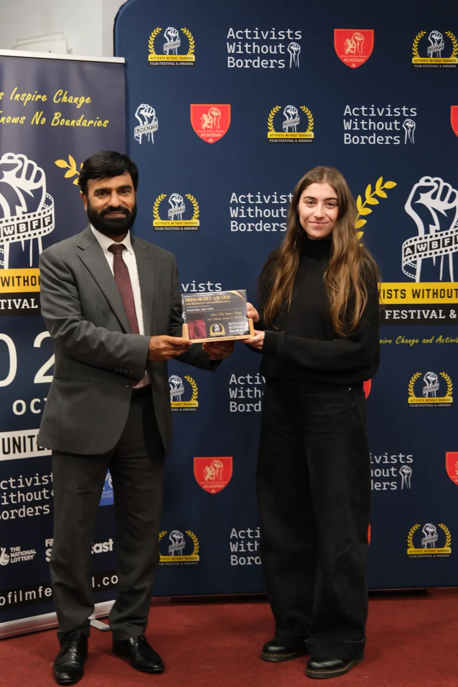 A man in a dark suit and a young woman in black hold an Honorary Award plaque between them in front of the festival backdrop