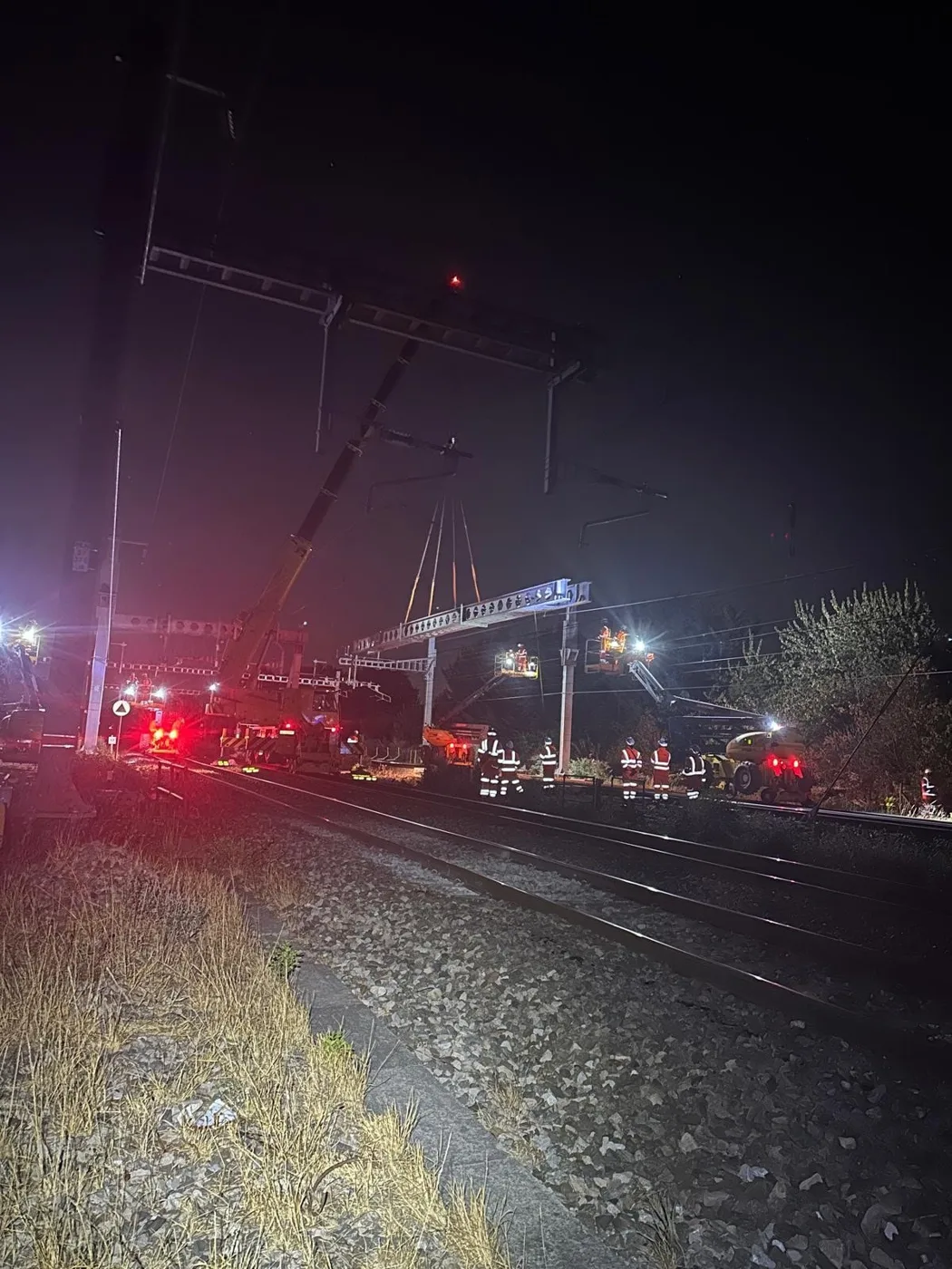 Engineers in orange high-visibility clothing work beside the track at night under floodlights, with a crane lifting a steel gantry for the overhead wires.