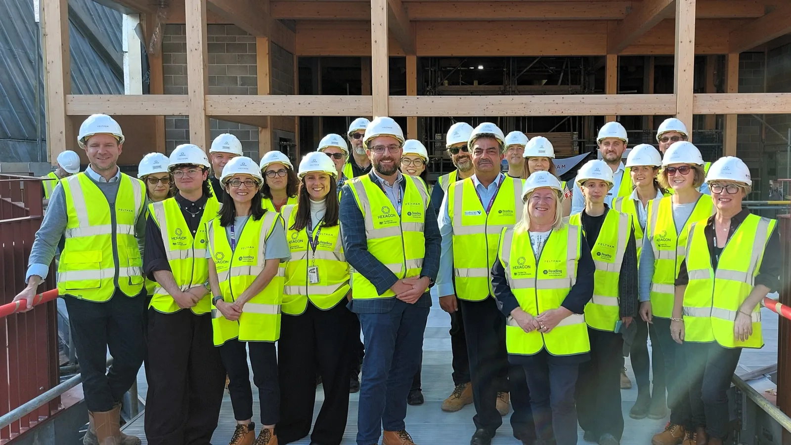 A group of about twenty people in white hard hats and yellow high-visibility vests standing on the new pedestrian walkway, with the timber frame of the Studio Theatre behind them.